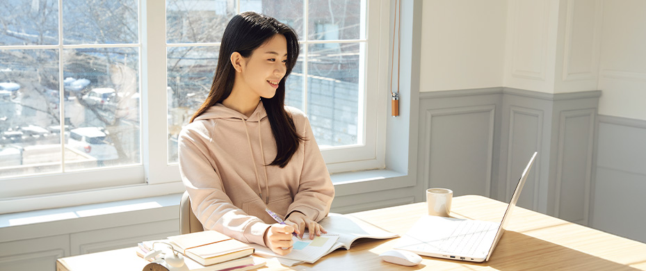 햇살 드는 창가 책상에서 분홍색 후드티 여성이 미소를 지으며 노트북과 공책으로 작업 중인 모습